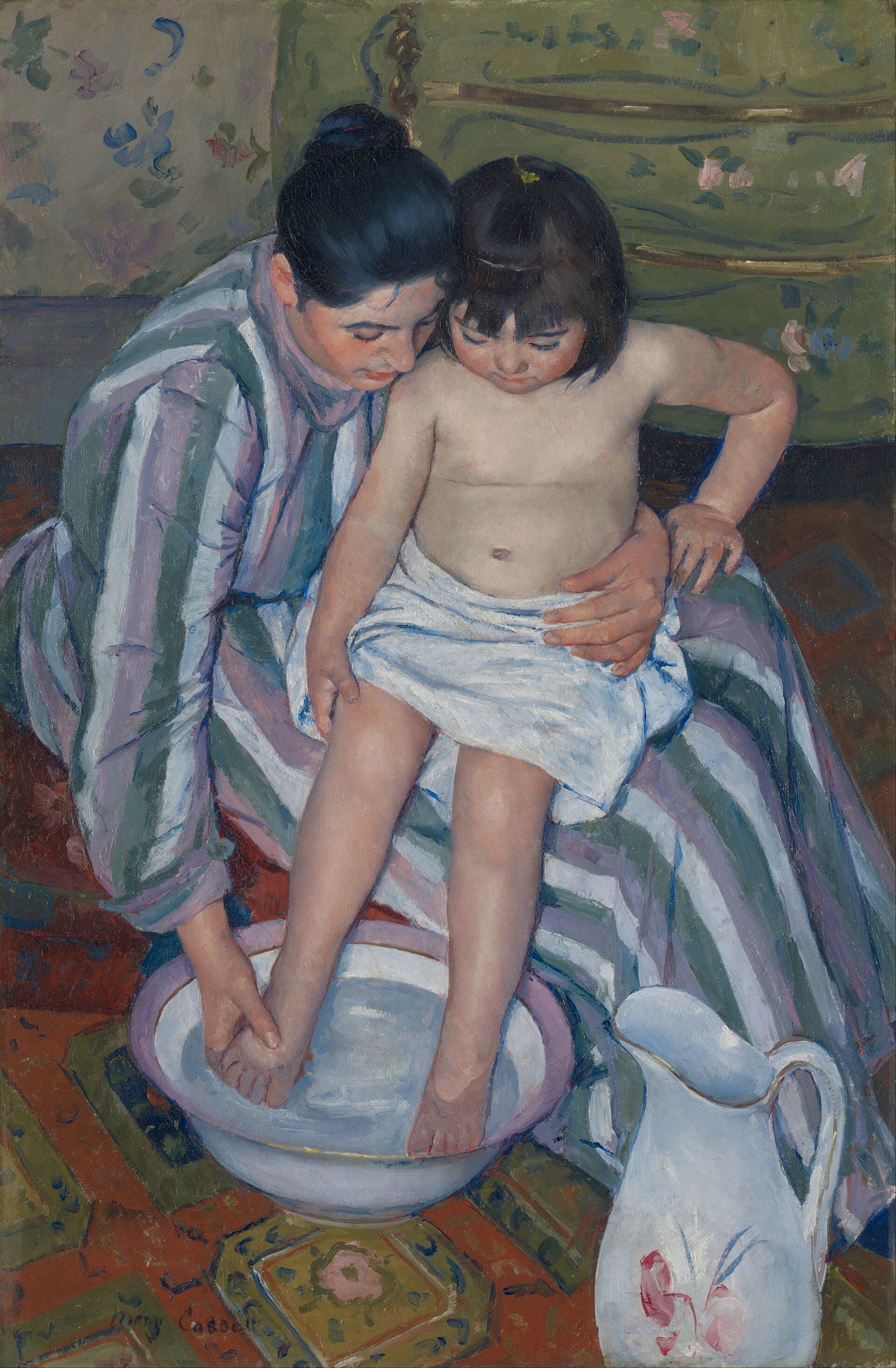 Seen from above, a woman in a striped dress holding a child on her lap and washing one foot in a basin, a patterned rug beneath them