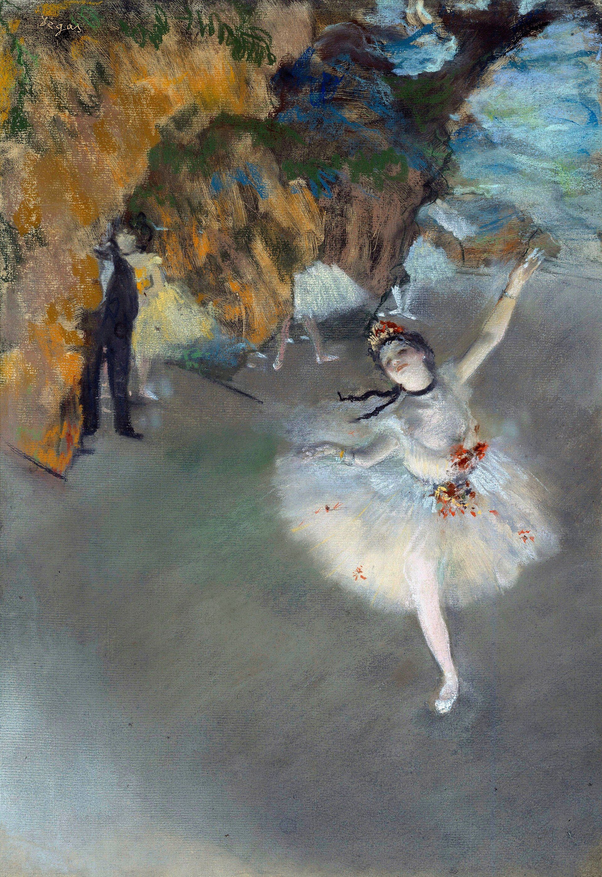A ballerina in white arriving at the front of a lit stage with one arm raised, painted scenery behind her and a man in black waiting in the wings