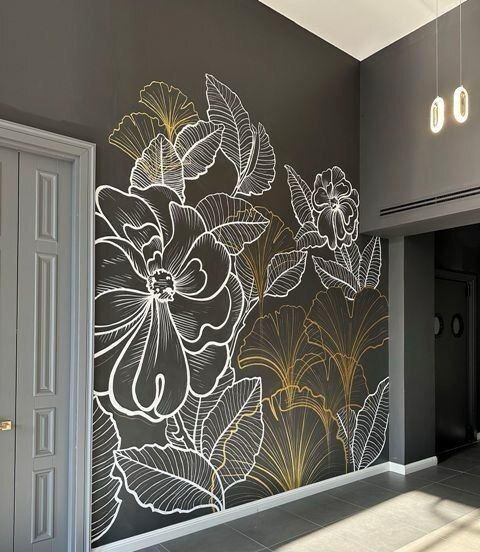 Dark grey hallway wall painted with oversized hibiscus, ginkgo and banana leaves drawn in white and gold outline, beside a panelled door