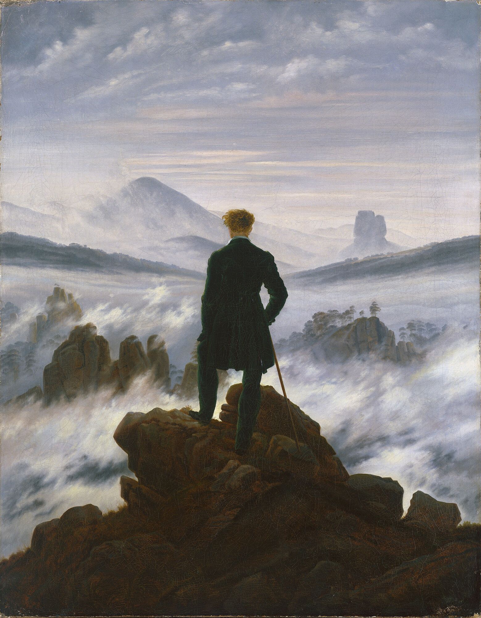 A man in a dark coat standing on a rock outcrop with his back turned, looking over cloud filling the valleys below