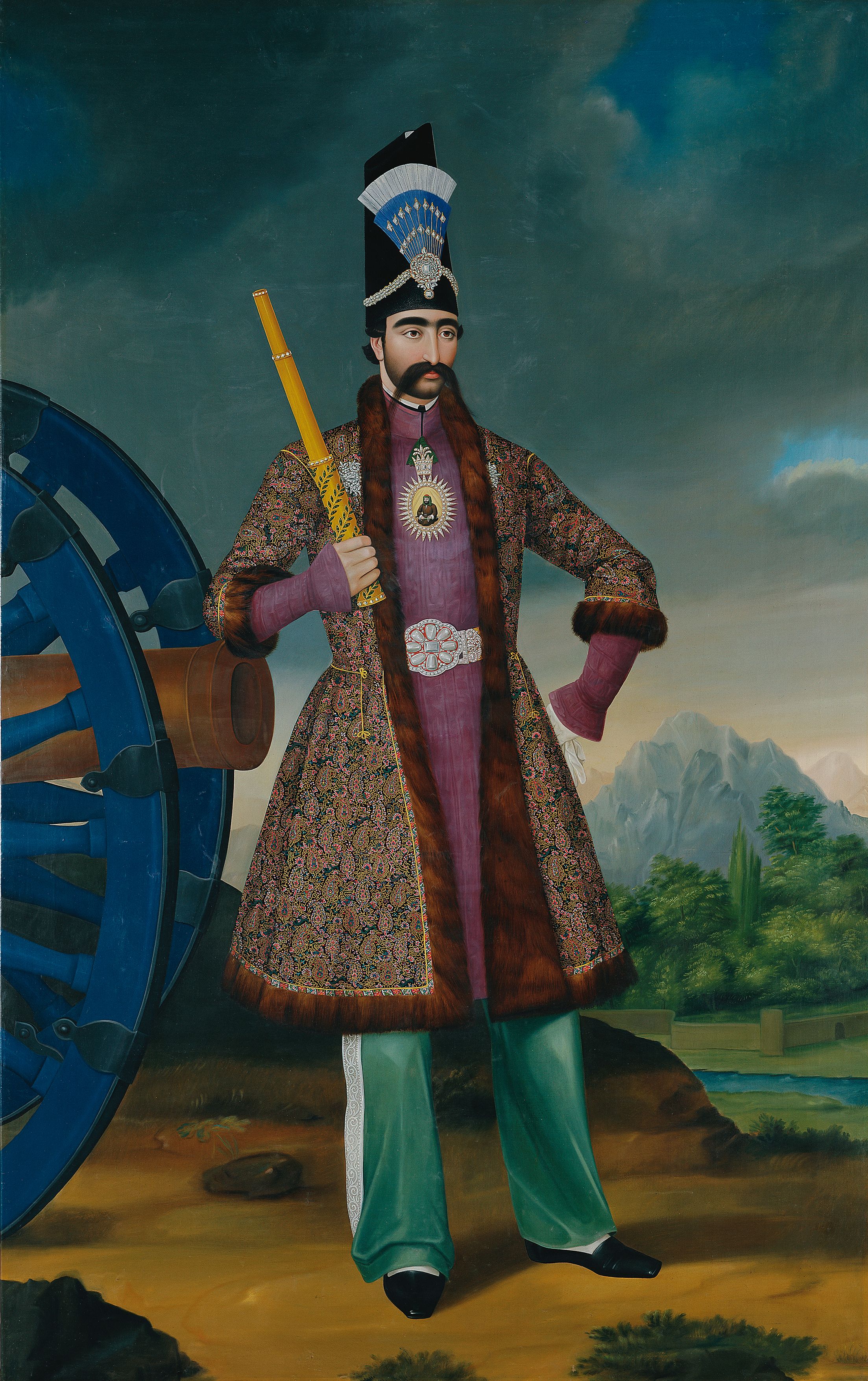 Full-length portrait of the Shah in a jewelled black hat and richly patterned brocade coat, standing beside a blue-and-brass cannon with mountains behind
