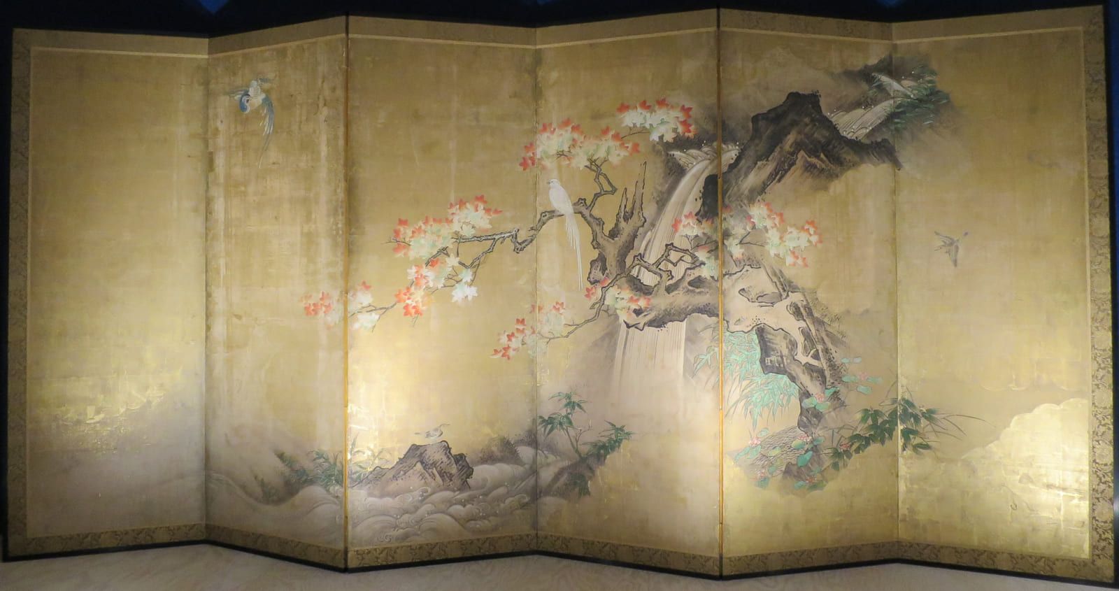 A gnarled maple leaning over a pale waterfall with small birds among its branches, on a six-panel gold screen