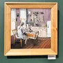 The same painting framed in pale wood, hung on a green gallery wall beside its label