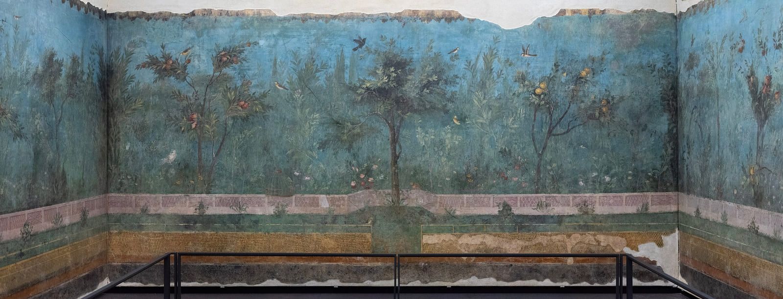 The end wall of a painted garden room, trees and shrubs banked behind a low fence under a wide band of blue