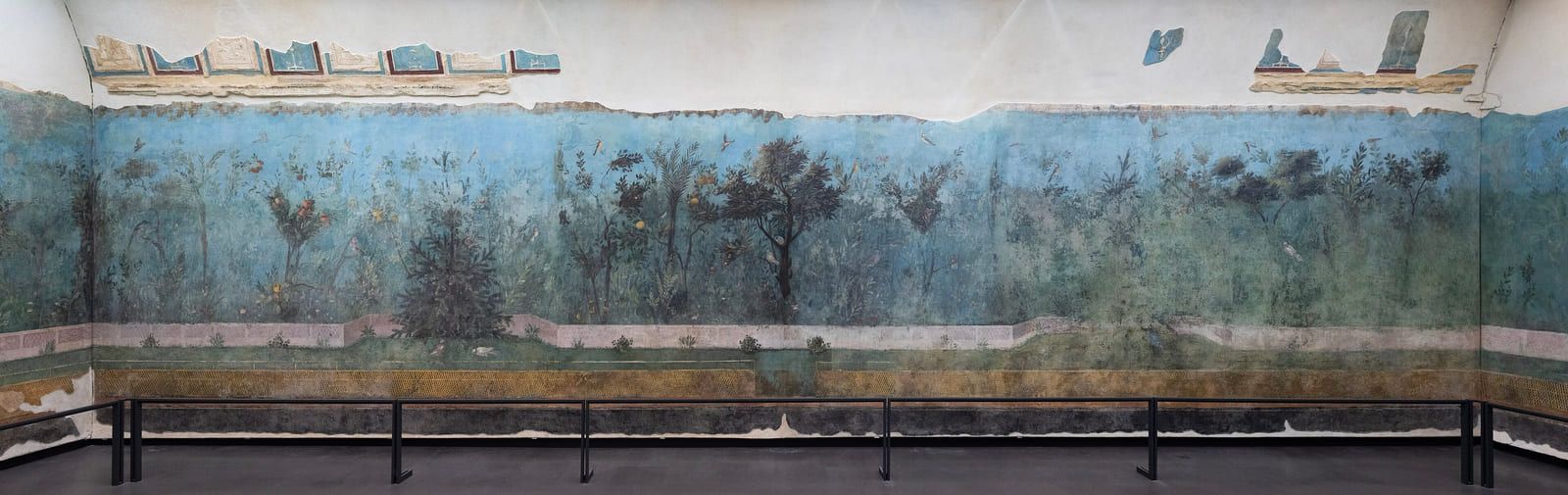 A very wide painted garden of dark trees against blue, running the full length of a wall above a low painted fence
