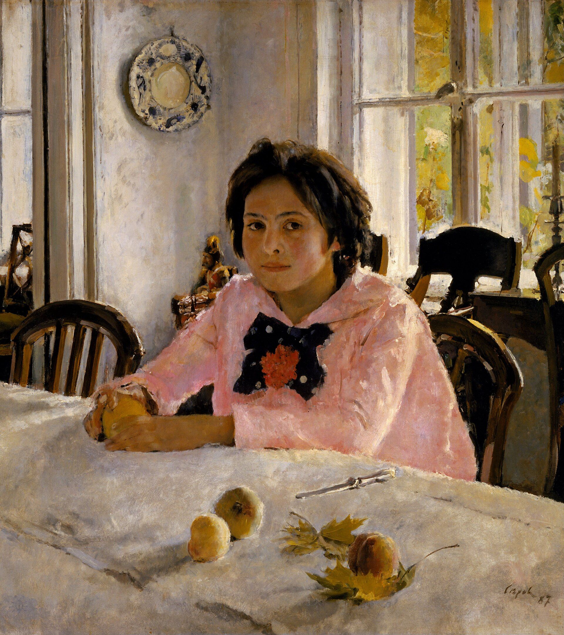 A dark-haired girl in a pink blouse with a black bow seated at a white cloth, peaches and a knife in front of her, tall windows behind