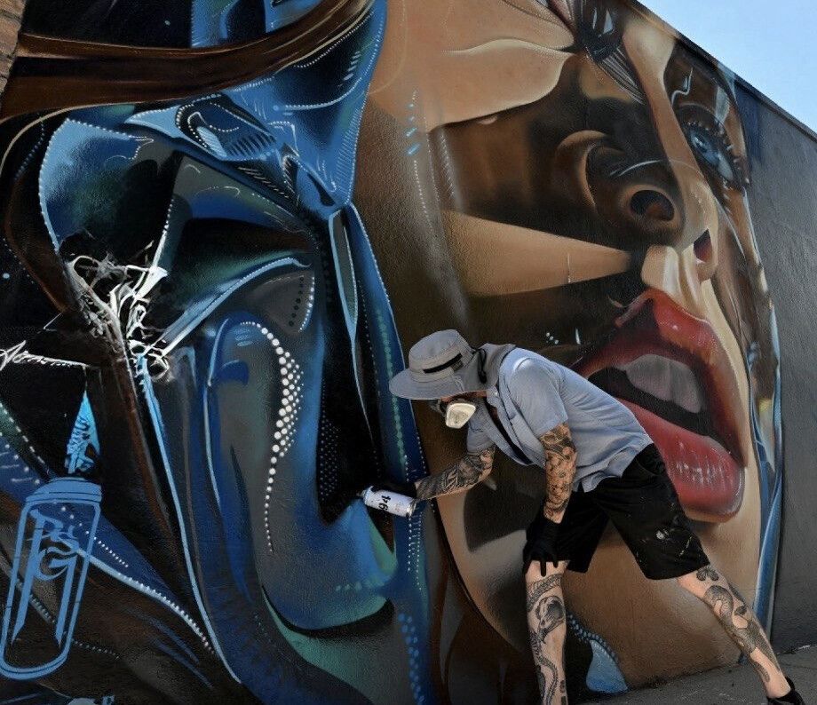 A woman's face in warm ochre beside folded blue drapery and lettering, sprayed along a long street wall, a painter at work at its foot for scale