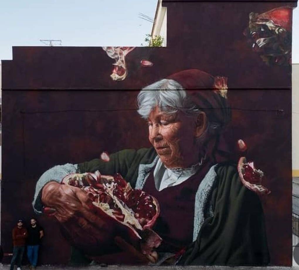 An elderly woman in a green coat and red headscarf holding a split pomegranate, painted the height of a windowless building on a deep red ground, seeds falling around her