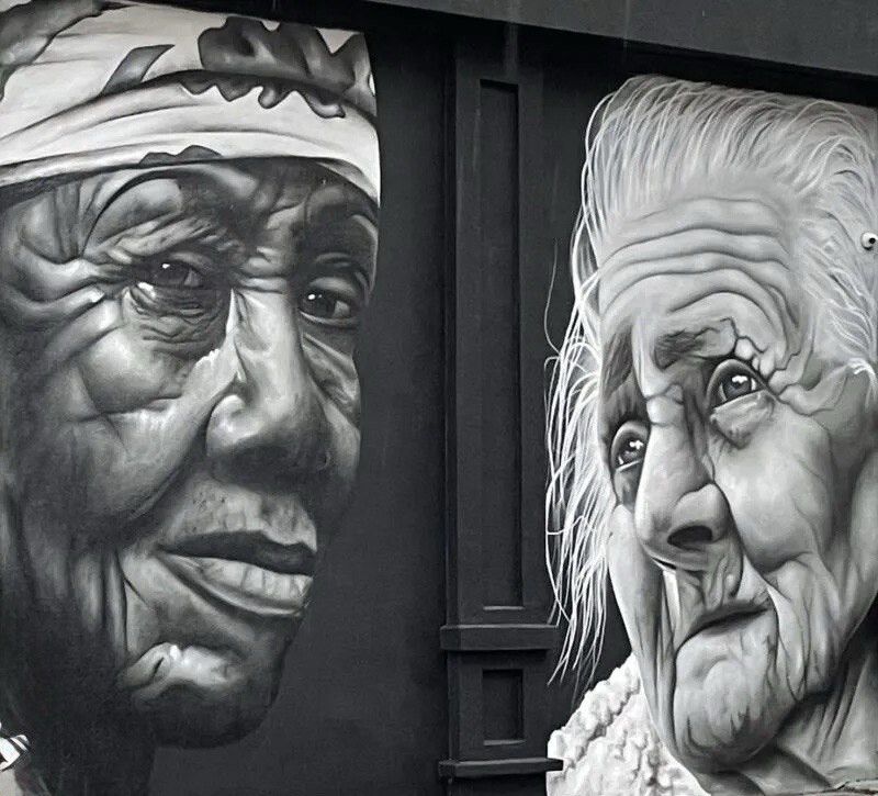 Two aged faces in grey and white on a black wall, one in a patterned headscarf, the other with loose white hair, worked in photographic detail