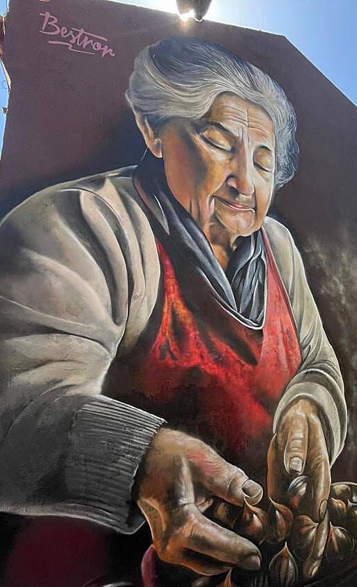 An elderly woman in a red apron bent over her cupped hands shelling walnuts, painted up a narrow gable end against open sky
