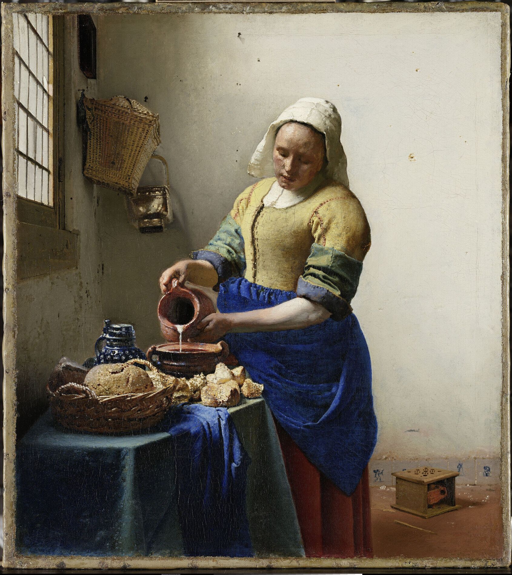 A servant in a yellow bodice and blue apron pouring milk from a jug at a table of bread, lit from a window at the left